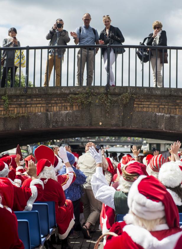 Eine Gruppe als Weihnachtsmänner verkleideter Menschen winkt von einem Boot aus Passanten auf einer Brücke zu.