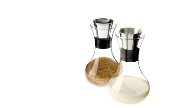 Zwei Glasbehälter, einer mit Zucker und einer mit Milch, stehen auf einem weißen Hintergrund.