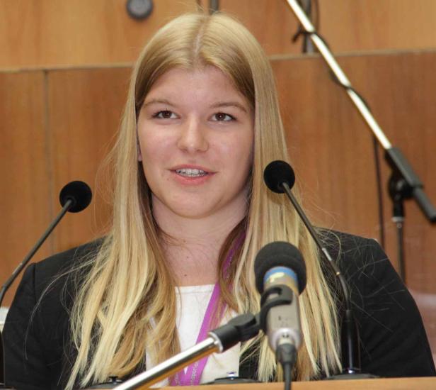 Eine junge Frau mit blonden Haaren spricht an einem Rednerpult mit mehreren Mikrofonen.