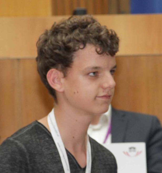 Ein junger Mann mit lockigem Haar und einem Namensschild um den Hals blickt nach rechts.