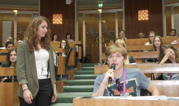 Ein junger Mann spricht in einem Hörsaal in ein Mikrofon, während eine Frau daneben steht.