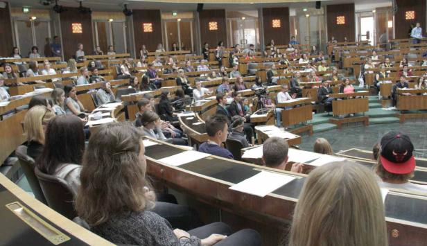 Zahlreiche Menschen sitzen in einem großen Hörsaal mit hölzernen Tischen und Stühlen.