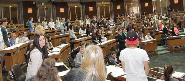 Eine Gruppe junger Leute sitzt in einem großen Saal mit Holzbänken.