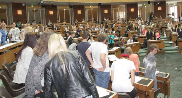 Viele junge Menschen sitzen in einem großen Saal mit Holzbänken.