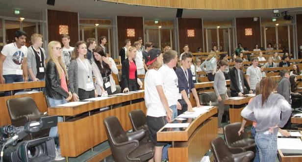 Eine Gruppe junger Leute steht in einem großen Saal mit Holzbänken und Tischen.