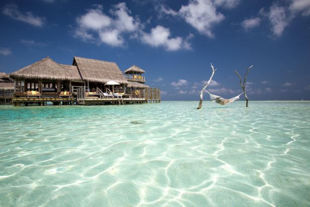 Ein strohgedecktes Resort steht auf Stelzen über dem türkisfarbenen Wasser, mit einer Hängematte im Vordergrund.