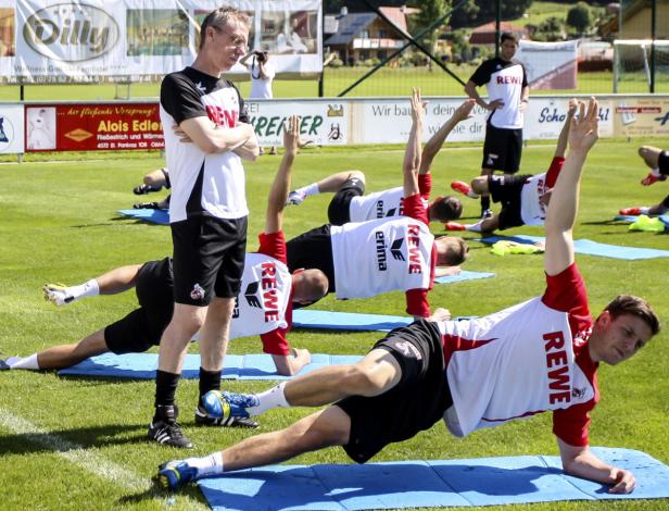 Eine Gruppe von Sportlern macht Dehnübungen auf blauen Matten im Freien, während ein Trainer zusieht.