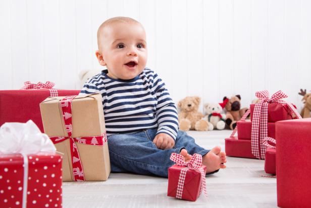 Ein Baby sitzt zwischen Weihnachtsgeschenken und Stofftieren.