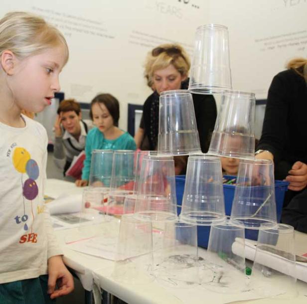 Ein Mädchen betrachtet einen Turm aus Plastikbechern auf einem Tisch.