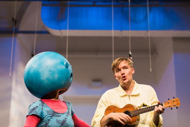 Ein Mann spielt Ukulele neben einer Person mit einer blauen Kugel als Kopf.