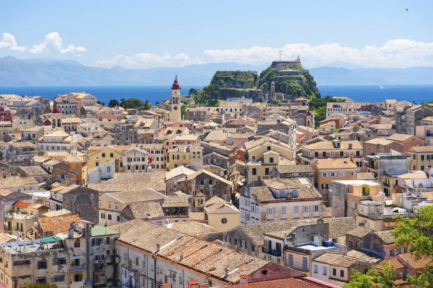 Die Altstadt von Korfu Stadt aus der Luft. Orangefarbene Ziegeldächer, ein Kirchturm und ein Hügel mit großem Kreuz.