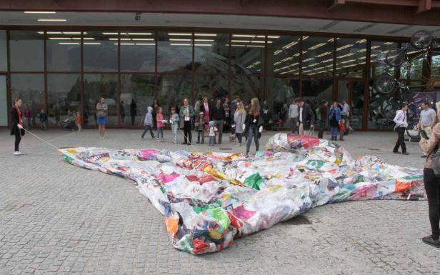 Eine große, bunte Skulptur aus recycelten Plastiktüten vor einem Gebäude mit vielen Menschen im Hintergrund.