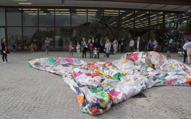 Eine große, bunte Skulptur aus recycelten Materialien liegt auf einem Platz vor einem Gebäude.