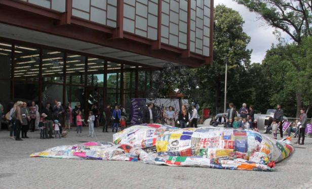 Eine Installation aus recycelten Materialien vor einem modernen Gebäude mit vielen Menschen.