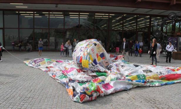 Eine große, bunte Skulptur aus recycelten Materialien liegt auf einem Platz vor einem Gebäude.