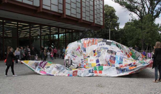 Eine große, mit Aufklebern bedeckte Skulptur vor einem Gebäude zieht die Aufmerksamkeit einer Menschenmenge auf sich.