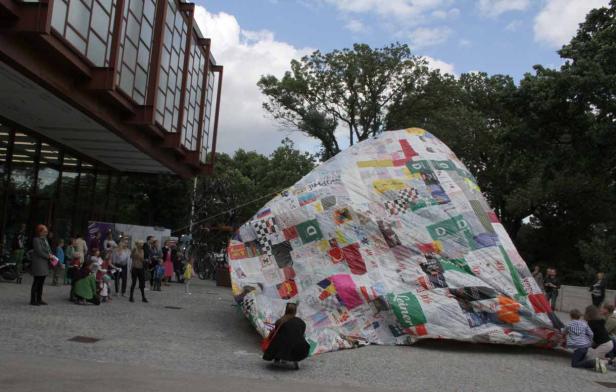 Eine große, patchworkartige Skulptur steht vor einem Gebäude, umgeben von Menschen.