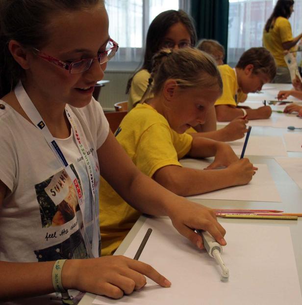 Eine Gruppe Kinder sitzt an Tischen und zeichnet mit Stiften und einer elektrischen Zahnbürste auf Papier.