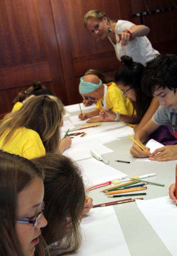 Eine Gruppe von Kindern sitzt an einem Tisch und zeichnet mit Buntstiften, während eine Lehrerin zusieht.