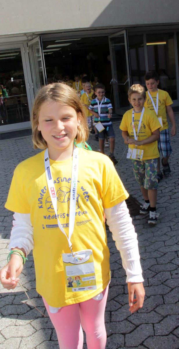 Eine Gruppe Kinder in gelben T-Shirts mit dem Aufdruck „Kinder Business Week“ verlässt ein Gebäude.