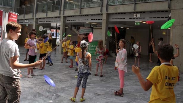 Eine Gruppe Kinder balanciert Teller auf Stäben in einem Innenraum.