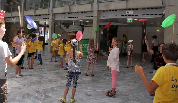 Eine Gruppe Kinder balanciert bunte Teller auf Stäben in einer Halle.