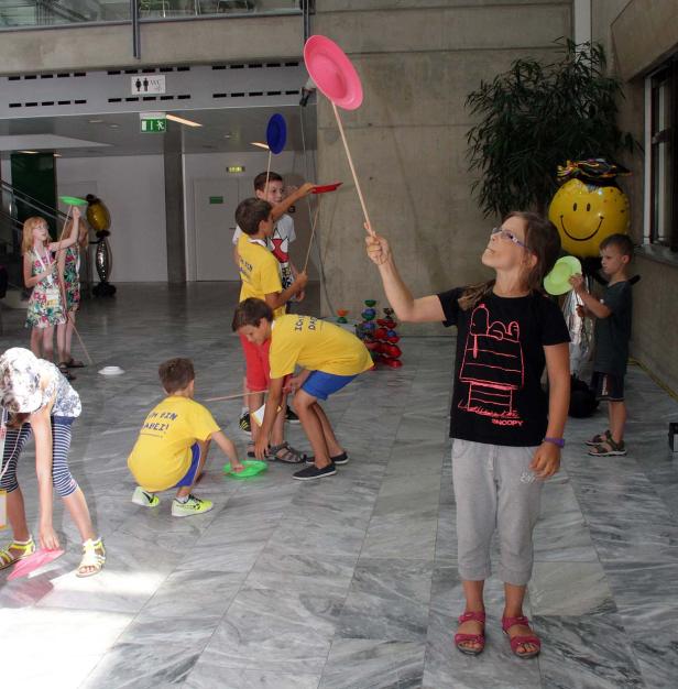 Eine Gruppe Kinder balanciert Pappteller auf Stäben in einem Innenraum.