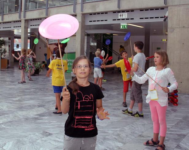 Eine Gruppe Kinder balanciert Teller auf Stäben in einem öffentlichen Gebäude.
