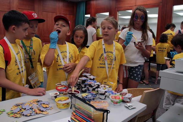 Eine Gruppe Kinder in gelben T-Shirts steht an einem Tisch mit Joghurt und Puzzlespielen.