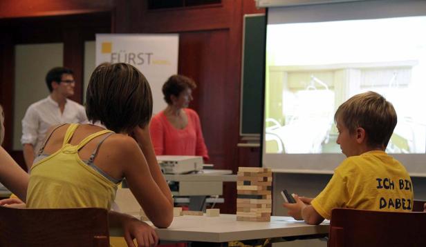 Kinder sitzen vor einer Leinwand und spielen mit Holzklötzen.