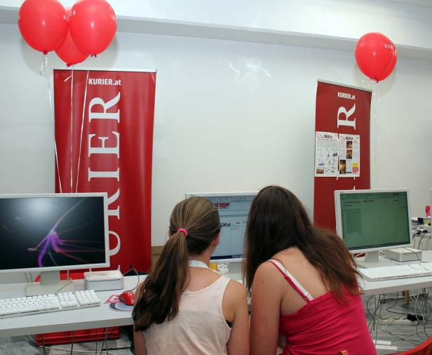 Zwei Frauen sitzen vor Computern mit „Kurier.at“-Bannern und roten Ballons im Hintergrund.