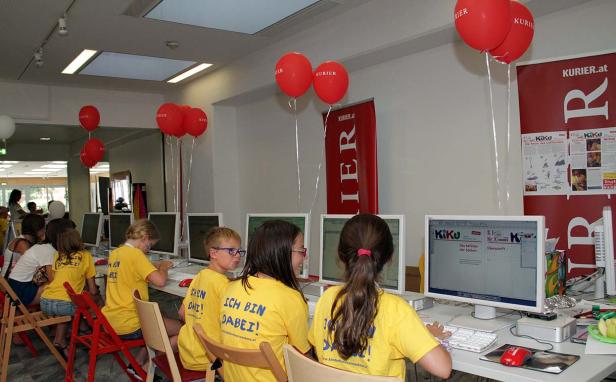 Kinder sitzen an Computern in einem Raum, dekoriert mit roten Luftballons und „Kurier“-Bannern.