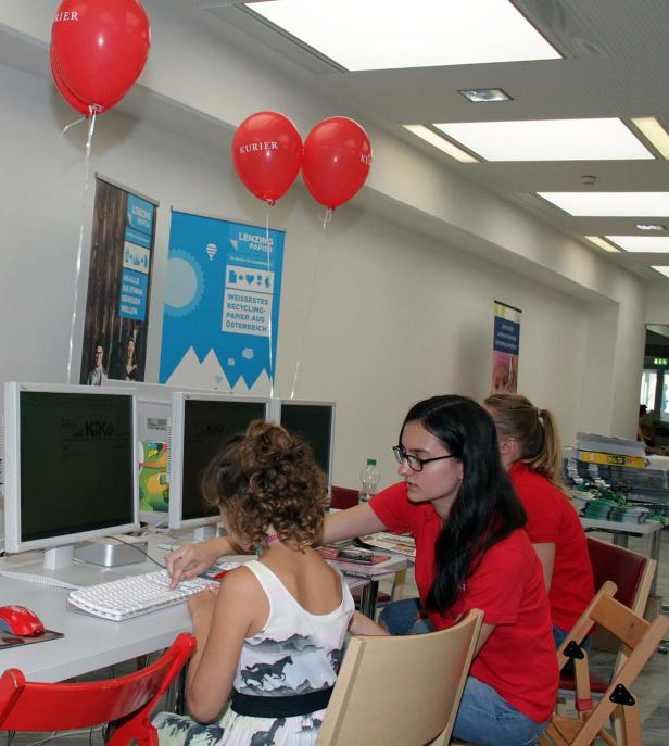Zwei junge Frauen sitzen an Computern unter roten „Kurier“-Luftballons.