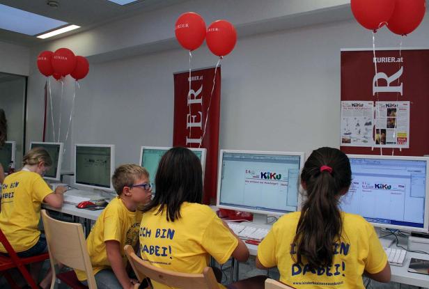 Kinder sitzen an Computern mit roten „Kurier“-Luftballons im Hintergrund.
