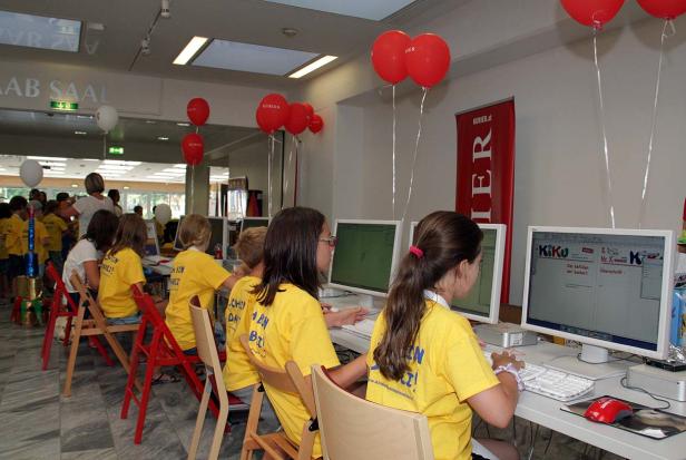 Kinder sitzen an Computern, geschmückt mit roten Luftballons, bei einer Veranstaltung des „Kurier“.