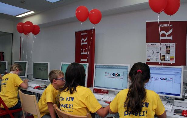 Kinder sitzen vor Computern mit roten Luftballons und Bannern des „Kurier“ im Raum.