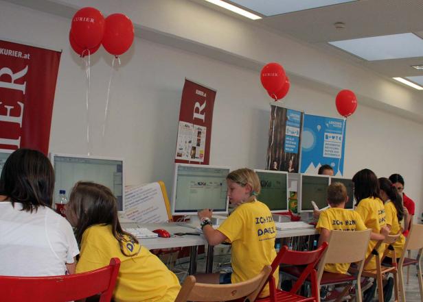 Kinder sitzen an Computern in einem Raum, dekoriert mit roten „Kurier“-Luftballons.