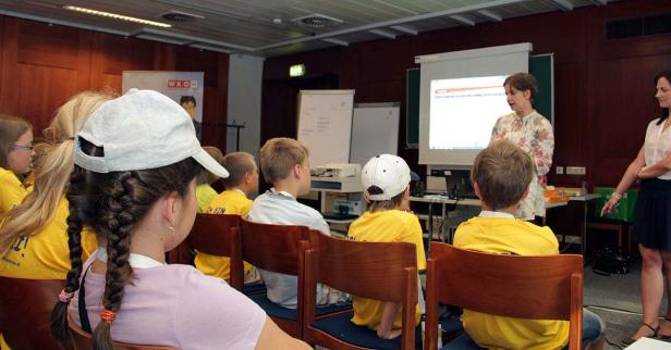Kinder sitzen in einem Raum und hören einer Präsentation zu.