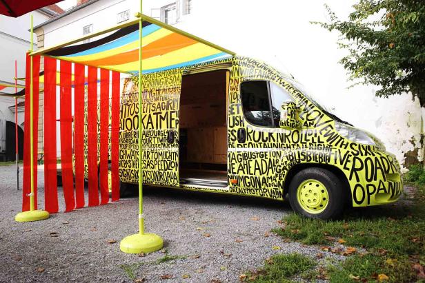 Ein gelber Lieferwagen mit schwarzer Schrift steht unter einem bunten Vordach.