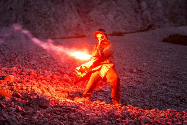 Eine Person in Pestdoktorkleidung hält eine rote Leuchtrakete in einer felsigen Landschaft.