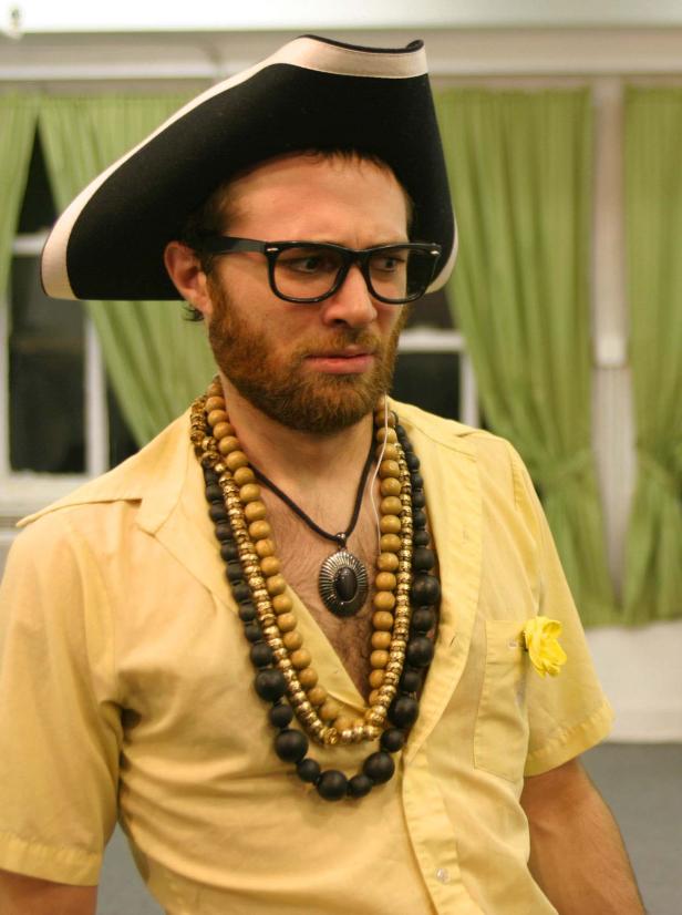 Ein Mann mit Bart, Brille und Dreispitz trägt mehrere Halsketten und ein gelbes Hemd.