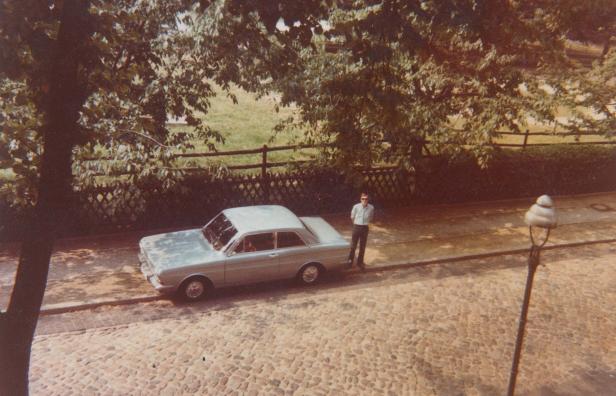 Ein Mann steht neben einem hellblauen Auto auf einem Kopfsteinpflasterweg.