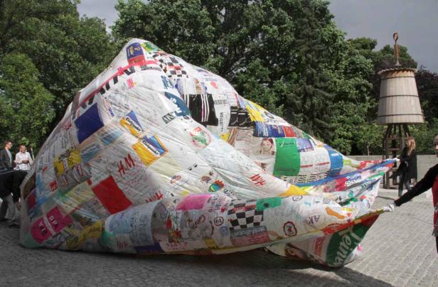 Eine große, mit Logos und Schriftzügen beklebte Struktur liegt auf einem Platz.