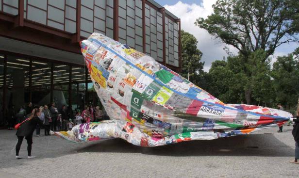 Eine große, walartige Skulptur aus recycelten Modezeitschriften steht vor einem Gebäude.