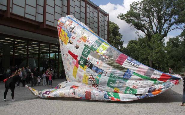 Eine große, walartige Skulptur aus recycelten Werbematerialien steht vor einem Gebäude.