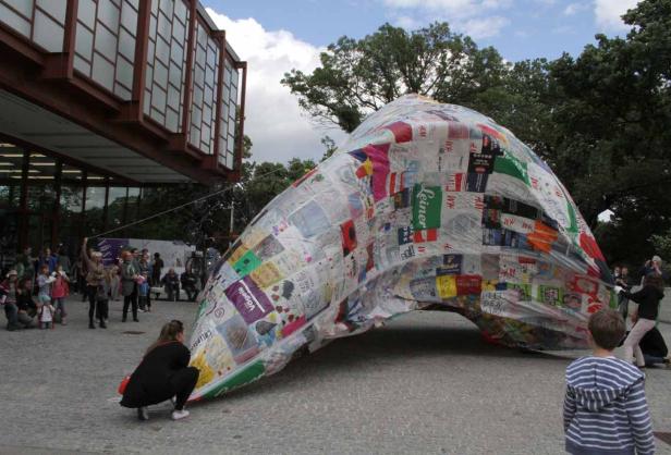 Eine große, bunte Skulptur aus recycelten Materialien steht im Freien vor einem Gebäude.