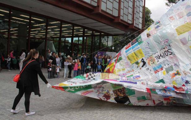 Eine Frau hält ein großes, mit Collagen bedecktes Objekt vor einem Gebäude, während eine Menschenmenge zusieht.