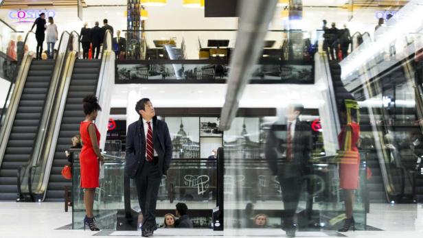 In einem Einkaufszentrum stehen eine Frau in einem roten Kleid und ein Mann im Anzug vor einer Rolltreppe.
