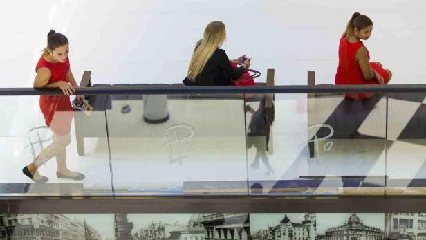 Drei Frauen in roten Kleidern sitzen und stehen auf einer Empore in einem Einkaufszentrum.