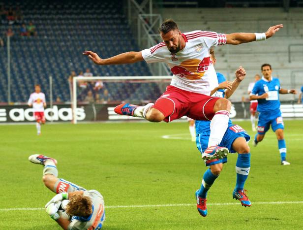 Ein Fußballspieler springt über einen am Boden liegenden Torwart während eines Spiels.